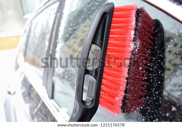 Red plastic brush cleaning snow. Shoveling snow from\
automobile with chrome details. Grey car outside covered with snow\
and ice. Frozen window glass with selective focus at the winter\
blizzard day.