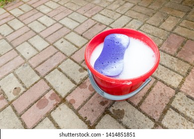 A red plastic bucket on cobblestone background with purple colored sponge. Cleaning theme.Household equipment at home interior background, spring-cleaning, tidying up, cleaning service concept - Powered by Shutterstock