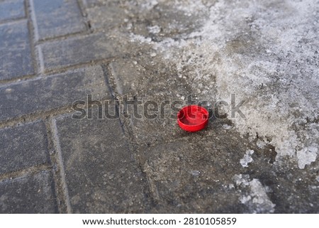 Red Plastic Cap Resting On Icy Urban Sidewalk With Melting Snow On Concrete Tiles Near Curb In Late Winter Daytime Closeup Of Discarded Small Object On Wet Pavement.