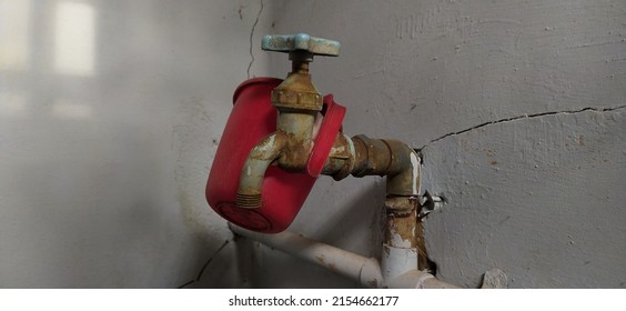 Red platic mug hanging on closed old vintage plastic water faucet tap pipe fixed on dirty and damaged indian bathroom toilet  wall. Closeup side view. unsanitary rural india village bathroom concept.