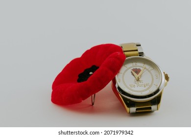 A red popy with black centre leaning on a gold and silver watch that says lest we forget, set to eleven  minutes past eleven o'clock