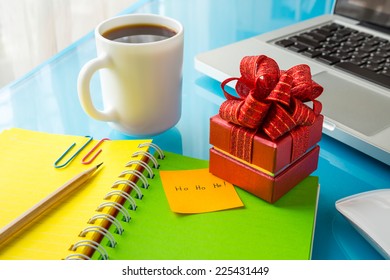 Red Present Box And Paper Note With Merry Christmas Message: Ho Ho Ho! Place On Office Table With Laptop, Colorful Diary And A Cup Of Coffee, Concept To Christmas Gift And Holiday Season