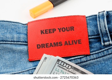 red rectangular label with the words keep your dreams alive rests on blue jeans with stacks of one hundred United States dollar bills, and a highlighter.