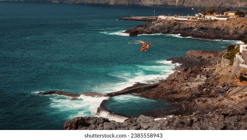 Red rescue helicopter flies over a stormy ocean and lifts a person on board near rocky coastline. - Powered by Shutterstock