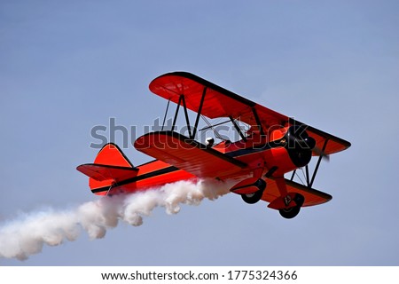 Red retro airplane (biplane) isolated on blue sky background. Vintage old red airplane & pilot flying in sky. View of red airplane & white smoke. Biplane condensation trail, smoky effect after plane