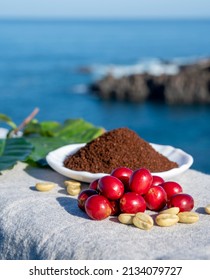 Red ripe arabica coffee berries, green coffee beands, leaves and roasted ground coffee in bowl close up and blue ocean view