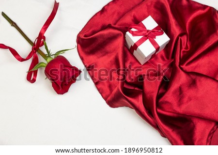 Red rose with red ribbon and red satin fabric with gif box on white bed with pillow. Valentine’s day or holiday romantic background