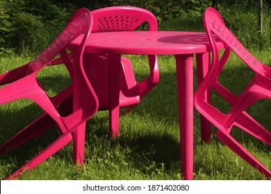 A Red Round Plastic Table With Chairs Leaning Against It, Standing On Green Grass Among The Greenery.