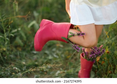 Red Rubber Boots On The Feet Of A Girl With A Bouquet Of Wild Flowers. Flowers In A Boot, Summer Time. Summer, Freedom, Nature, Countryside, Green Grass In The Field