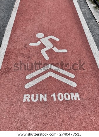 A red running track section marked with white graphics, featuring a pictogram of a runner, directional chevron arrows, and the text ‘RUN 100M.