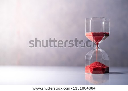 Red sand running through the shape of modern hourglass on white table.Time passing and running out of time. Urgency countdown timer for business deadline concept with copy space