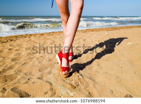 Red sandals on the legs of a girl standing on the beach sand