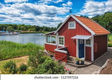 Red Scandinavian Wooden House On The Lake With Beautiful Blue Sky.