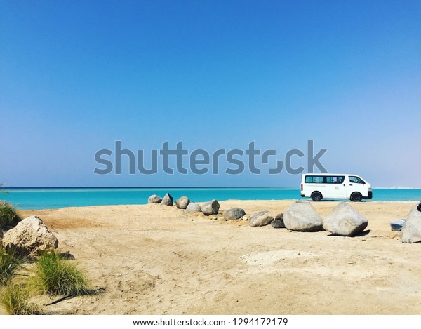 The Red Sea off the West coast of Saudi Arabia\
with sand and rocks leading to the waters edge and a white van\
parked in the distance.