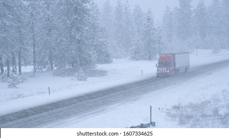 Red semi truck hauls a heavy cargo container across the state of Washington and through a snowstorm. Freight lorry navigates the slippery country road in the low visibility during an intense blizzard.
