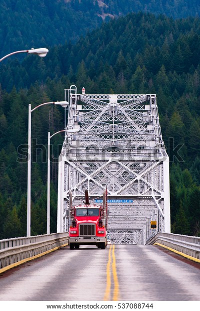 Red semi truck rides on the road with a\
yellow stripe dividing across the silver metal bridge on the\
background mountains covered with dense\
forest