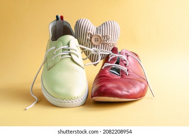 A Red Shabby Male And Female Shoe With A Heart Between Them On A Yellow Background. The Concept Is A Relationship Between An Older Man And A Younger Woman. Leather Shoes.