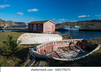 A red shed sits on the edge of a bank near the water. There's a blue fishing boat in the background and a couple of old wooden boats grounded with worn red paint. There's mountains in the background.