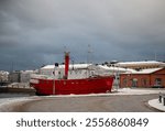 Red ship docked on a snowy waterfront with festive lights, surrounded by historic buildings under a cloudy sky