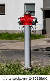 A red and silver fire hydrant located on a gray pole in front of a white building. The fire hydrant has a yellow nozzle and a black cap. There is a green inspection tag attached to the pole.