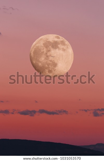 red sky during sunset\
with a big moon in the full moon phase and the silhouette of a\
small plane. image of nature without people. image of the moon with\
tinting in red.