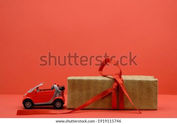 Red small car toy near the unpacked box with a red\
ribbon. Red background. Gift, surprise concept. Buy car concept.\
Copy space.