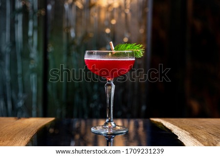 A red sour cocktail in a coupe glass, decorated with a fir branch on the table in a bar