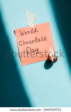 Red square paper handwritten note world chocolate day with one small candy egg glued with adhesive tape on a blue background with hard shadows, close-up side view. World chocolate day concept.