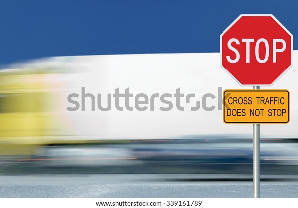 Red stop road sign, motion blurred truck vehicle\
traffic in background, regulatory warning signage octagon, white\
octagonal frame, metallic pole post, yellow cross traffic does not\
stop text signage
