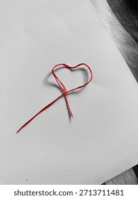 Red string carefully formed into shape of heart, lying on white piece of paper. Simple, love and affection. Background softly blurred, emphasizing heart. Handmade card, decorative for design