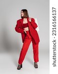 red suit, bold makeup, tattooed young woman with brunette short hair and nose piercing holding shopping bags and standing on grey background, youth culture, fashionable outfit, consumerism