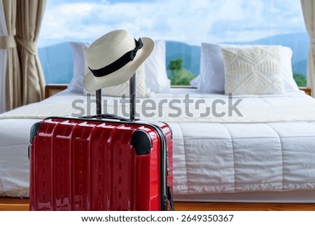 Red suitcase with straw hat placed in front of a cozy prepared fresh bed with mountain view and cloud sky in natural sun light. Ready for a peaceful vacation in nature. Rainy season.