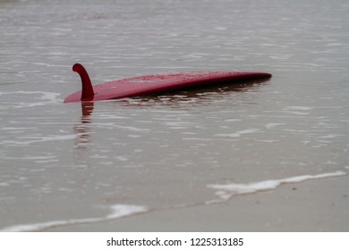 Red surfboard lying upside down on the sand at the edge of the shore at the beach.  