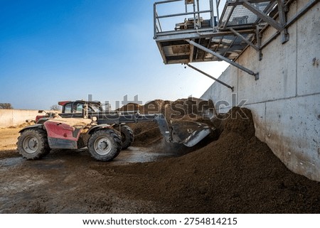 A red telehandler moves a pile of solid digestate in a modern biogas facility. The image highlights agricultural automation and renewable energy management under a clear blue sky, showing industrial waste processing and sustainability.