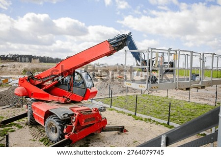 Red telescopic handler elevates a work platform over green crops at a construction site, with a clear blue sky and clouds visible in the background
