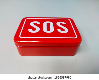 A Red Tin Container Of SOS Containing Some Essential Equipment When In An Emergency. SOS Survival Emergency Gear Self Help Outdoor Camping Hiking Tools Box Kit Set In White Background. Random Focus.