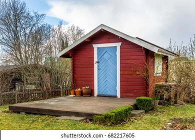 Red Tiny House Or Shed At A Plot Of An Allotment. Early Spring Time. Small Red Shack With The Blue Door, White Frames, Flat Wooden Terrace And Flowers. Hut Painted In Traditional Swedish Red Color.