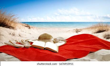 Red Towel On Beach With Book And Hat And Ocean Landscape. 