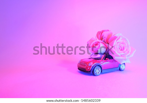 Red toy car delivering bouquet of pink rose flowers\
on trendy color neon background. Place for text. February 14 card,\
Valentine\'s day. Birhday. 8 March, Women\'s Day. St.Petersburg\
Russia 16 jul 2019