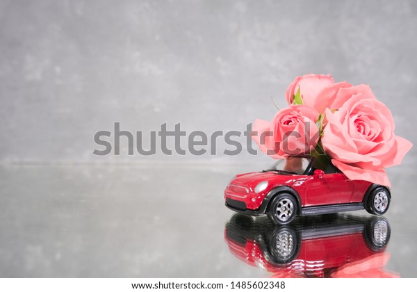Red toy car delivering bouquet of pink rose flowers\
on grey background. Mirror reflection. Place for text. February 14\
card, Valentine day. Flower delivery Women Day. St.Petersburg\
Russia 16 jul 2019