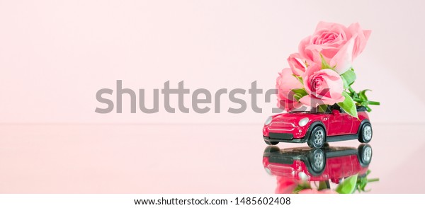 Red toy car delivering bouquet of pink rose flowers\
on pink background. Banner. Place for text. Mirror reflection.\
Valentine. Flower delivery. 8 March, Women\'s Day. St.Petersburg\
Russia 16 jul 2019.