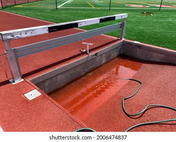 A Red Track And Field Steeplechase Pit Is Being Filled With Water By A Green Hose So Track Runners Can Jump Into It During Their Running Race.