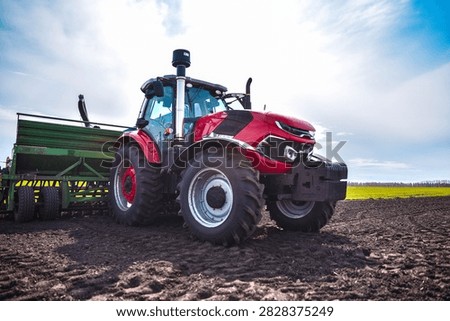 Red tractor pulling a modern seeder across a field during the planting season. Realistic agricultural scene showing active fieldwork and modern farming machinery.