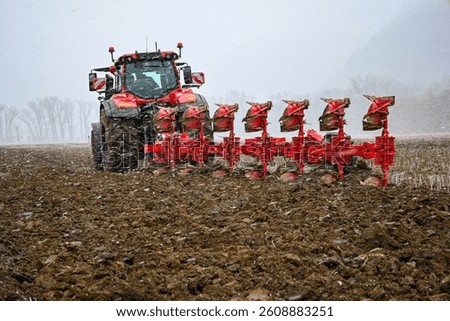 Red tractor pulling plow in field during snowfall, preparing soil for cultivation.