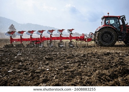 Red tractor pulling plow in field during snowfall, preparing soil for cultivation.