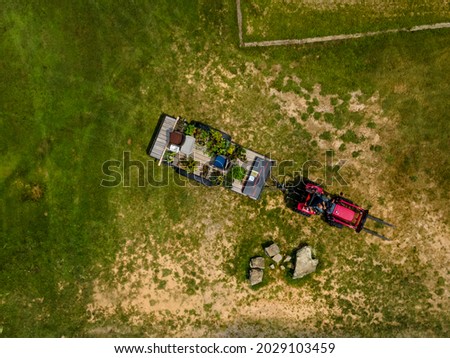 A red tractor pulls a trailor with plants on it through a patchy green grass field on a sunny clear summer day.  Aerial perspective looking down as the photograph was shot from an drone.