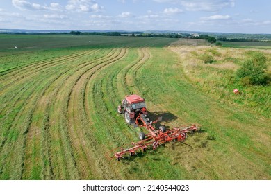 A red tractor rakes the mown grass for drying. Modern equipment on the field. Preparations of fodder grasses for hay. Top view. - Shutterstock ID 2140544023