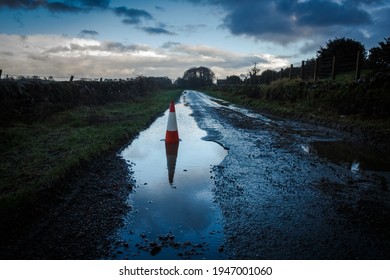 A Red Traffic Cone Reflecting A Pot Hole Containing A Pool Of Water On A Road, On A Cloudy Evening At Sunset