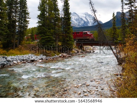 Red Train passing through a river bridge with the Canadian Rocky Mountains as Background.