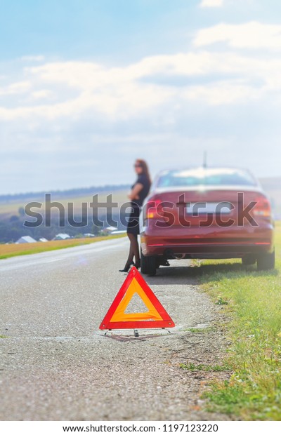 A red triangle an emergency stop sign\
against a blurred woman, the driver stands near her car and waits\
for help on the highway on a warm summer\
day.
